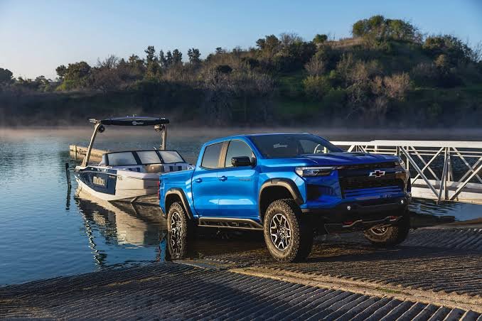 A bright blue Chevy Colorado (with 7,700 lbs max towing capacity) is shown towing a white boat down a lake slipway, demonstrating the chevy colorado towing capacity in a real-world scenario.