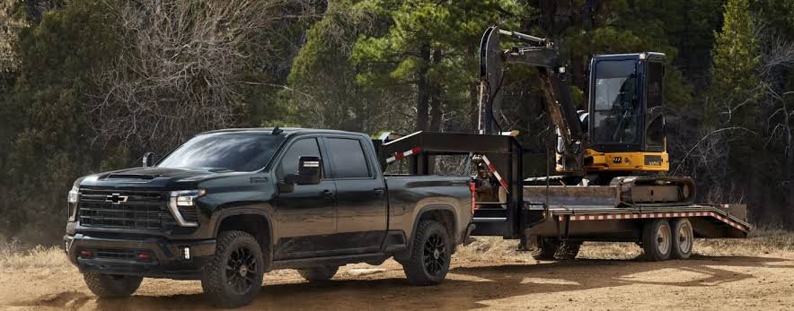 A black Chevy Silverado 1500 towing a flatbed trailer loaded with a heavy yellow mini excavator along a wooded dirt trail, demonstrating its impressive chevy silverado 1500 towing capacity.