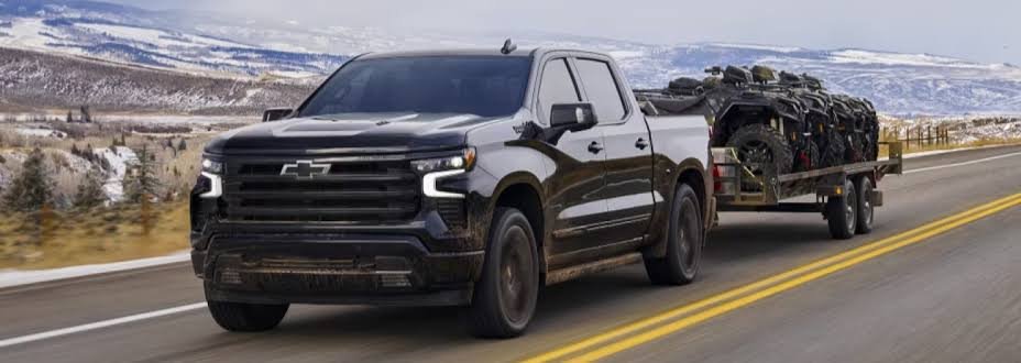 A black Chevy Silverado 1500 towing a trailer loaded with off-road vehicles along a snowy highway, showcasing its robust chevy silverado 1500 towing capacity.