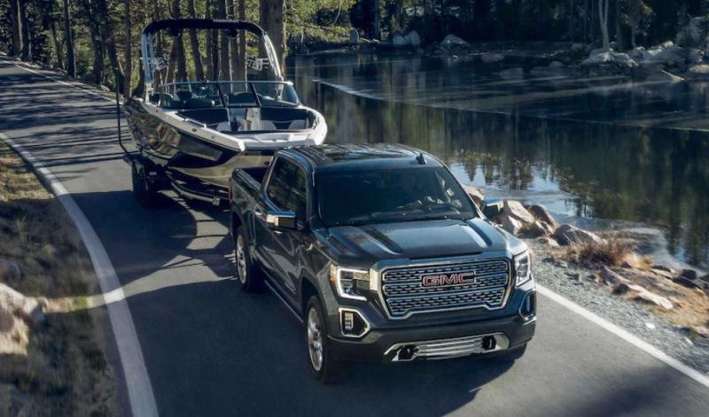 This image shows a dark GMC Sierra 1500 pickup truck effortlessly hauling a white boat on a trailer along a scenic road next to a calm lake, perfectly illustrating the vehicle's gmc sierra 1500 towing capacity.
