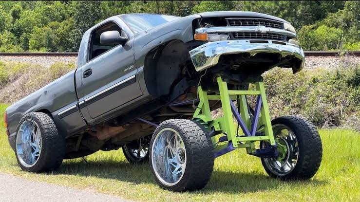 A grey squatted truck parked on grass, featuring an extreme front lift supported by custom green and purple tubular suspension brackets and large polished wheels.