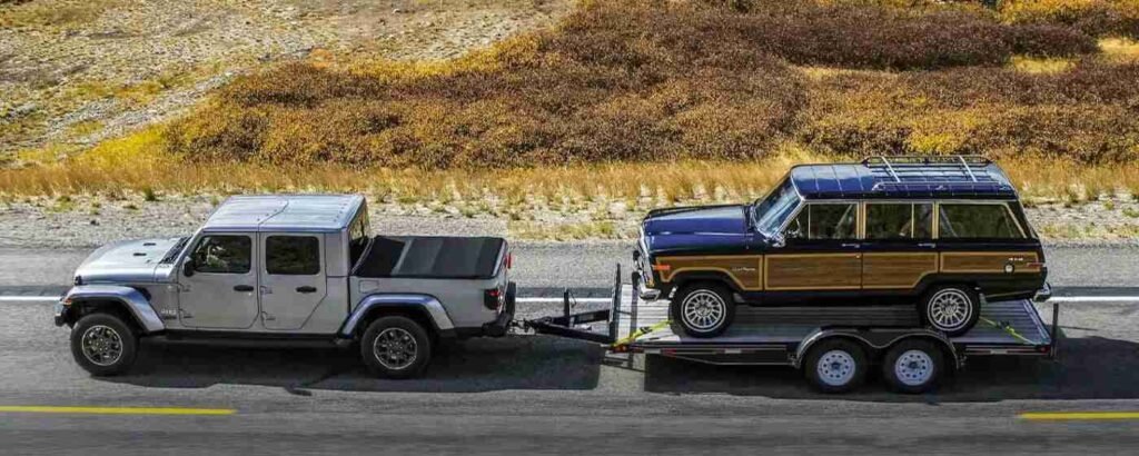 A silver hardtop Jeep Gladiator showcases its impressive jeep gladiator towing capacity while towing a classic wood-paneled Jeep Wagoneer on a flatbed trailer through autumn mountain scenery.