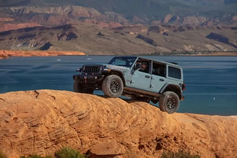 Gray off-road Jeep Wrangler climbing rocky terrain beside a blue lake, demonstrating the rugged frame quality related to jeep wrangler towing capacity.