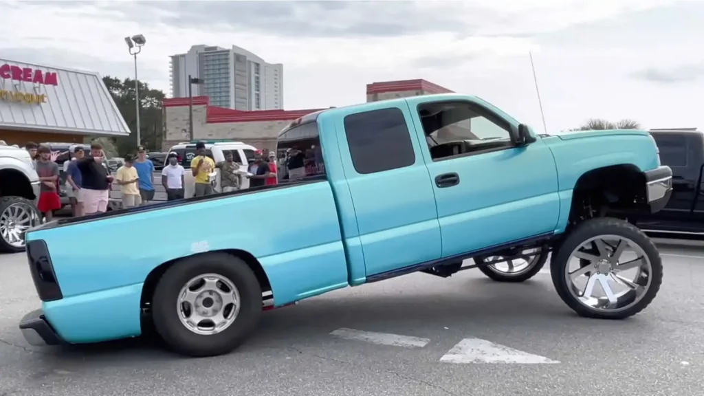 A light blue squatted truck with a heavily lifted front end and oversized front wheels parked at a car meet, with a crowd of spectators and buildings in the background.