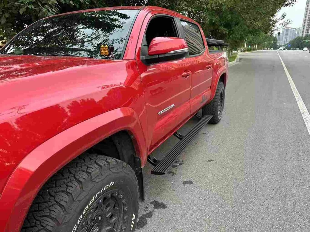 Side view of a red Toyota Tacoma pickup truck parked on a street, equipped with black aluminum alloy Power Running Board Materials, showcasing the extended step.