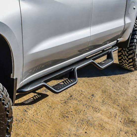 Black nerf bars with drop steps mounted on a silver pickup truck, parked on a dirt trail.