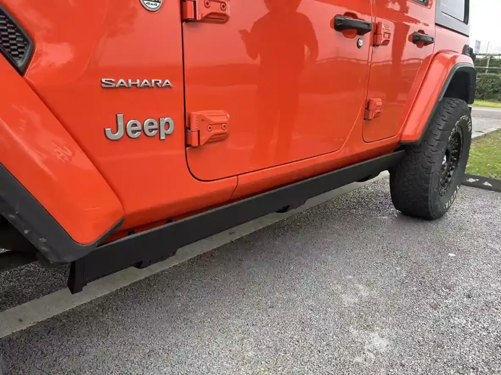 A side view of an orange Jeep Wrangler Sahara showing its black power running boards and the ground clearance beneath the vehicle.