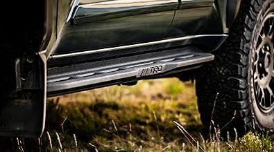 Close-up view of heavy-duty black running boards installed on a lifted truck, featuring a Raptor logo, parked on grassy terrain