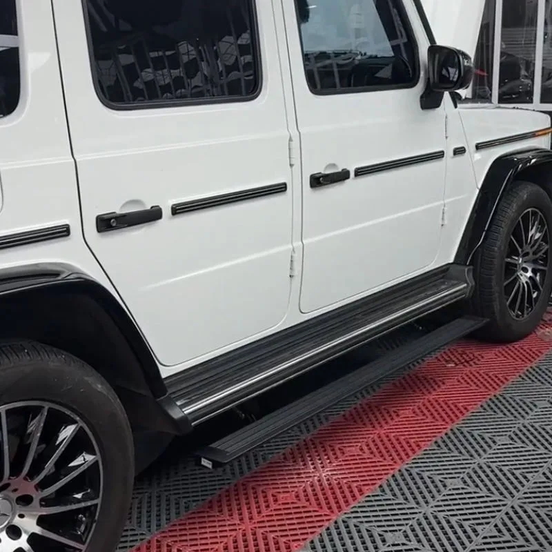 Side profile of a white Mercedes-Benz G550 showing the fully deployed Mercedes-Benz G550 Electric Running Boards resting above a red and black patterned floor mat.