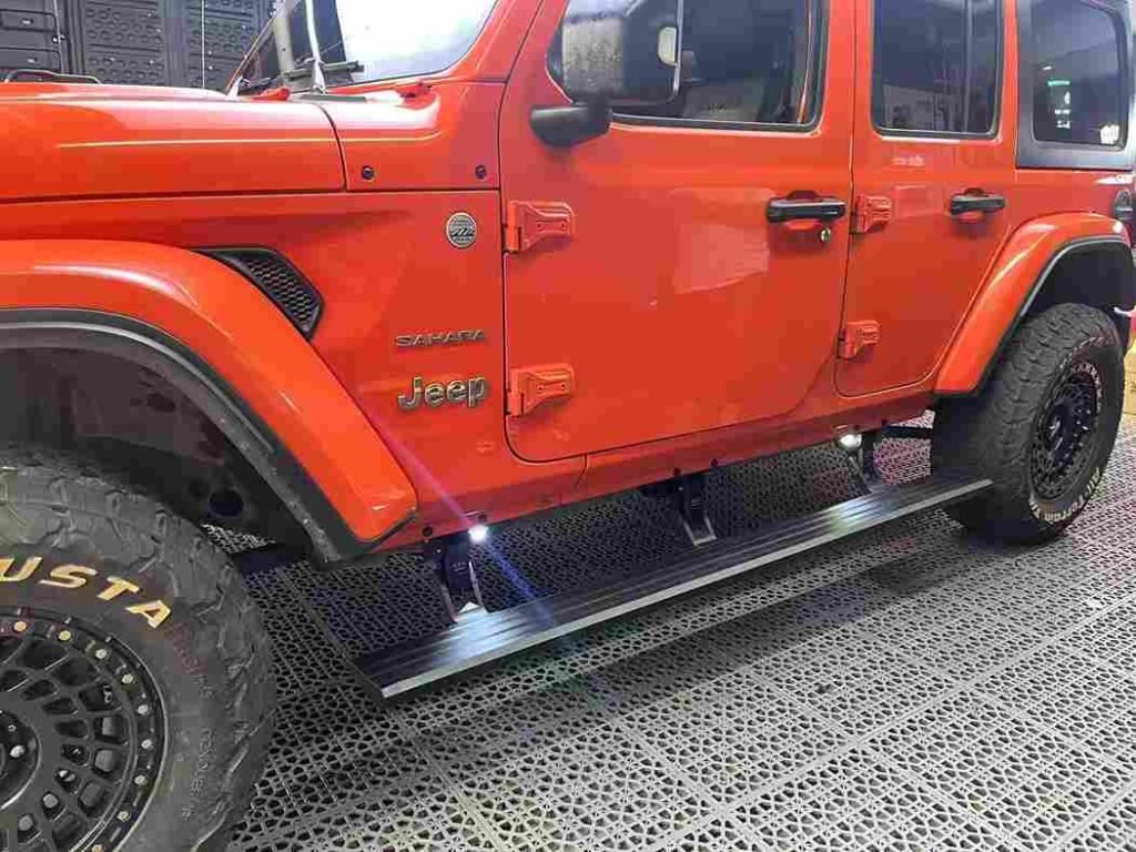 An orange Jeep Sahara featuring Electric Running Boards from the Expand 4x4 Shop Product Range. The retractable side steps are deployed and equipped with integrated LED spotlights illuminating the ground.