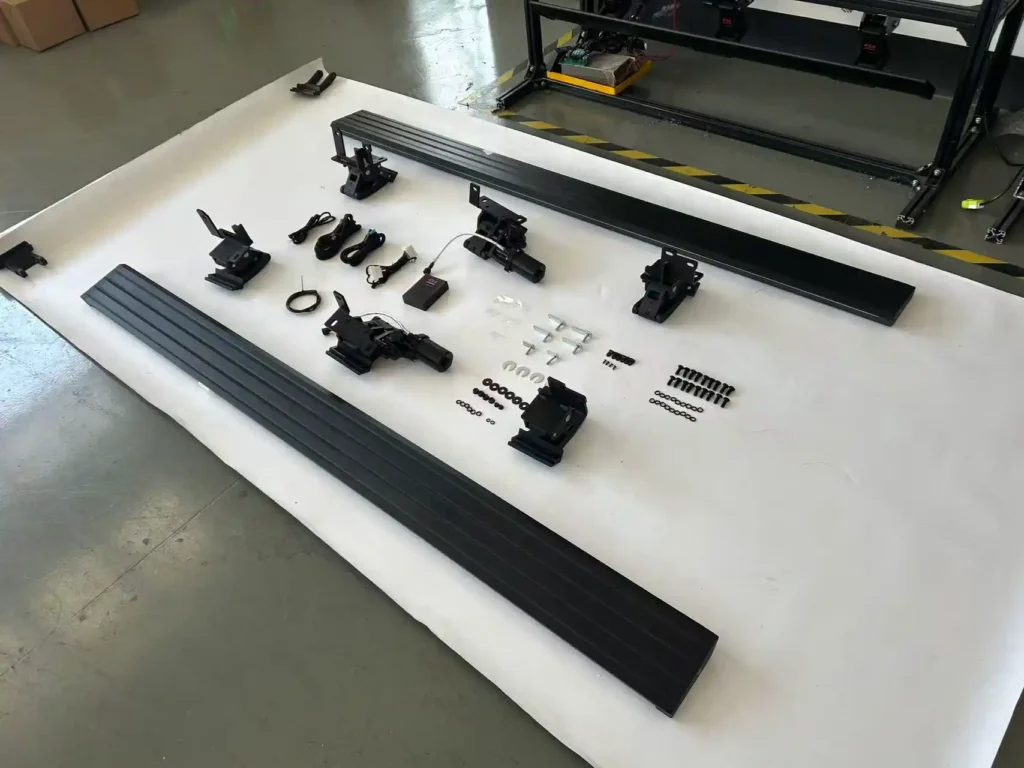 Exploded view of an Electric Running Board Sample kit laid out on a white surface, displaying two black step bars, electric drive motors, mounting brackets, a wiring harness, a control module, and various assembly hardware.