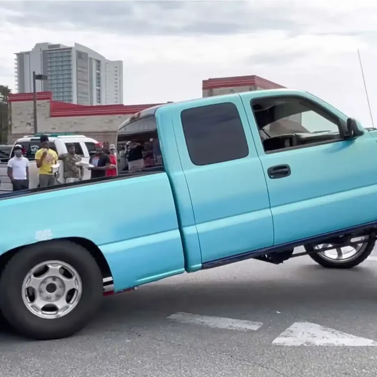 A light blue squatted truck with a heavily lifted front end and oversized front wheels parked at a car meet, with a crowd of spectators and buildings in the background.